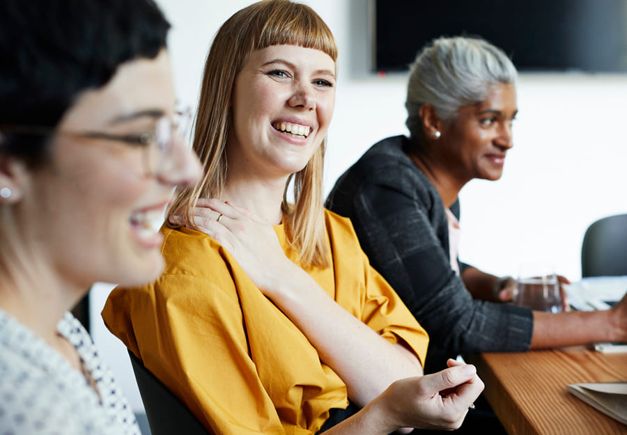 Three women sit at a table, all smiling. The woman in the centre is in focus - she wears a bright yellow shirt and is smiling broadly as she rests her left hand on her right shoulder comfortably. 