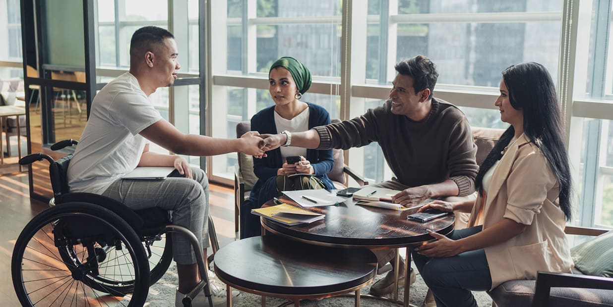 A diverse group of individuals gather around a table and engage in conversation. There are notepads and pens on the table. Two men shake hands across the table, one of whom is in a wheelchair. 