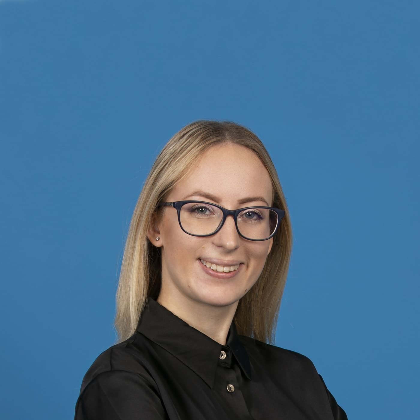 Young blonde woman wearing glasses and smiling into the camera in front of blue background.
