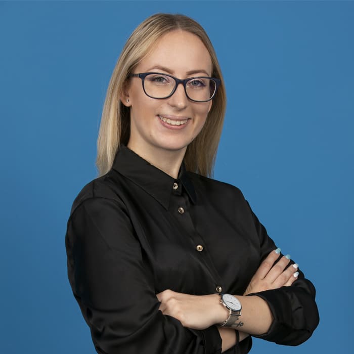 Young blonde woman wearing glasses and smiling into the camera in front of blue background.