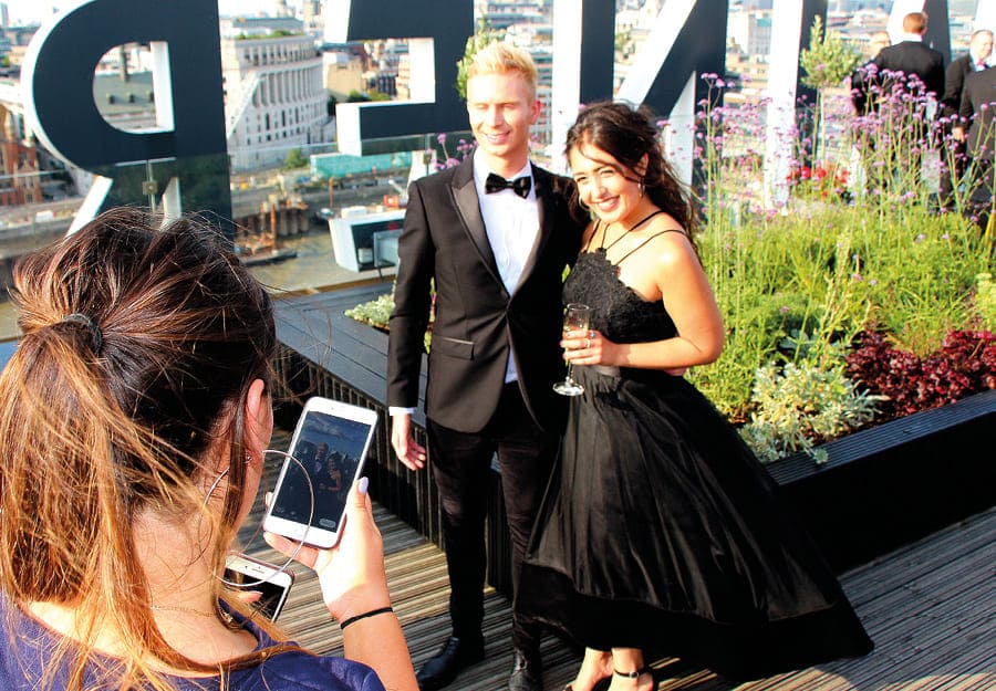 A young woman takes a photo of an elegant young couple in a rooftop bar